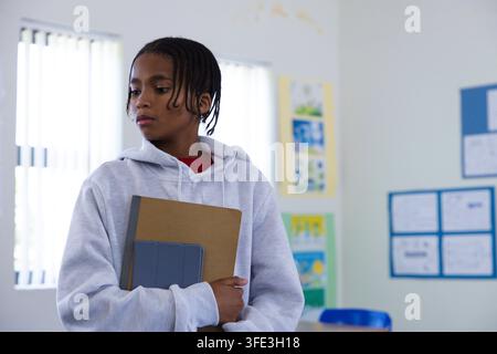 Étudiant adolescent portant un sweat à capuche gris debout à gauche à l'école tenant un cahier marron et une étui pour tablette Banque D'Images