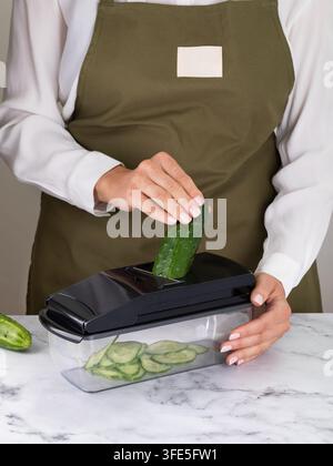 Femme coupant un concombre frais en fines tranches à l'aide d'une mandoline manuelle avec récipient transparent sur une table en marbre Banque D'Images