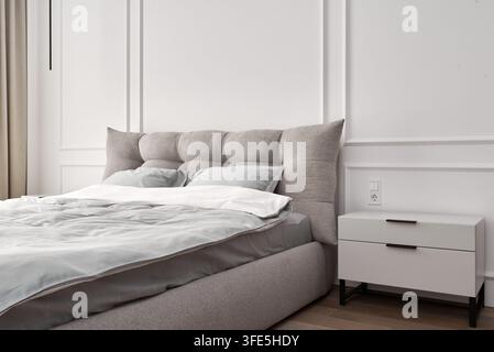 Intérieur de chambre moderne propre et lumineux avec une table de chevet blanche, un lit gris rembourré, une prise de courant, une conception de moulure murale et des tons beiges neutres. Coz Banque D'Images