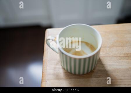 Une tasse à café rayée vide avec des résidus de mousse séchée repose sur une table en bois clair, une armoire de cuisine sombre est flouément floue en arrière-plan derrière elle Banque D'Images