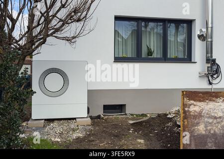 Une nouvelle pompe à chaleur air-sol se trouve à côté d'un bâtiment moderne. La pompe est placée sur du gravier près d'une fenêtre avec des rideaux. Les travaux de construction sont en cours aro Banque D'Images
