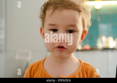 Garçon sérieux avec une expression réfléchie regarde l'appareil photo dans un portrait neutre. Tout-petit calme avec une attitude composée devant un fond simple à la maison. Enfant silencieux dans un moment de contemplation enfantine Banque D'Images
