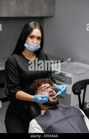 Dentiste femme portant un masque et des gants effectuant un bilan dentaire sur un patient masculin dans une clinique moderne. Se concentrer sur l'hygiène bucco-dentaire Banque D'Images