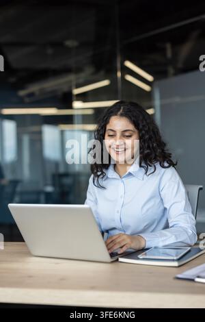 Jeune femme d'affaires souriante travaillant sur un ordinateur portable, tapant sur le clavier et regardant l'écran, reflétant la productivité et le bonheur dans un environnement professionnel de bureau Banque D'Images