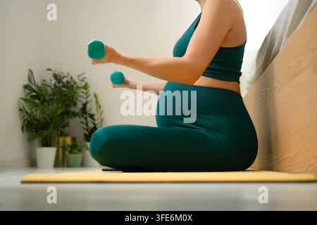 Femme enceinte sur tapis de yoga tenant des haltères légers pour des exercices prénataux. Mère en attente effectuant un entraînement sûr pendant la grossesse. Future maman maintenant sa forme avec un entraînement en force doux dans la chambre Banque D'Images
