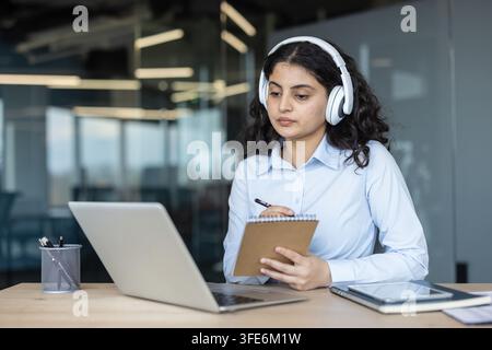 Jeune femme avec des écouteurs sur la tête prenant des notes dans un cahier tout en travaillant ou en étudiant en ligne sur son ordinateur portable à un bureau dans un environnement de bureau moderne Banque D'Images
