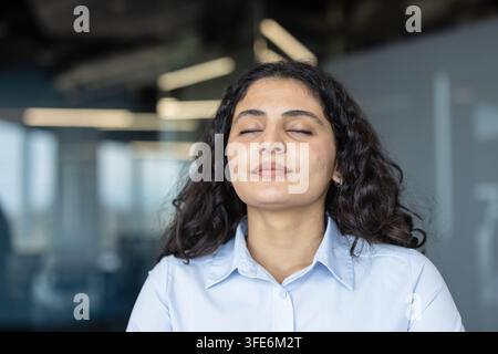 Jeune femme fermant les yeux, trouvant le calme et la paix grâce à une respiration profonde et à la pleine conscience dans un cadre de bureau d'entreprise, promouvant le soulagement du stress et le bien-être mental au travail Banque D'Images