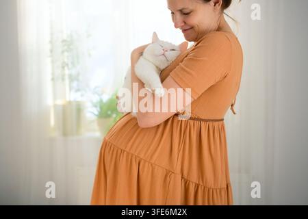 Une femme enceinte tient dans ses bras son chat blanc bien-aimé. La mère enceinte qui embrasse son compagnon animal présente un moment chaleureux et tendre de lien affectueux et de bonheur paisible à la maison. Banque D'Images