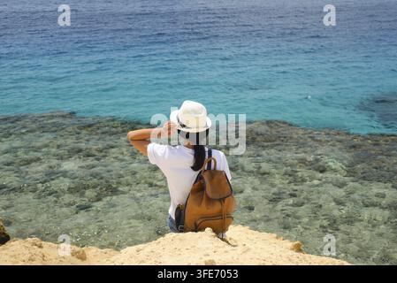 Voyageur solitaire avec chapeau et sac à dos assis sur une falaise rocheuse surplombant le paysage marin Banque D'Images