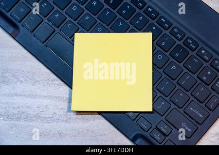 Vue par-dessus d'une note adhésive jaune carrée placée sur un clavier d'ordinateur noir sur un fond de bureau en bois. Espace de copie pour texte ou maquette. Banque D'Images
