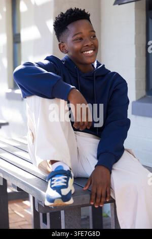 Adolescent afro-américain assis sur un banc près d'un mur de briques peint, portant un sweat à capuche bleu marine Banque D'Images