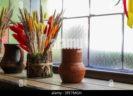 Arrangement de fleurs d'automne dans un vase en terre cuite sur le rebord de la fenêtre, scène chaleureuse et rustique avec des fleurs séchées chaudes et un support à fleurs en bois Banque D'Images