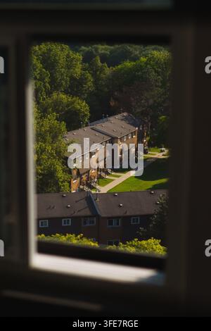 Vue en hauteur naturellement encadrée par une fenêtre donnant sur des maisons de ville en briques brunes de banlieue, des pelouses vertes et des arbres matures par un midi ensoleillé Banque D'Images