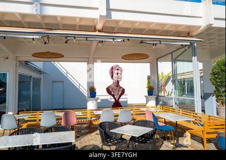 Une zone d'assise vide et baignée de soleil sur la terrasse d'un café contemporain, avec des tables, des chaises et une fresque murale frappante représentant une buste sur le mur Banque D'Images