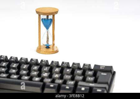 Sablier avec du sable bleu qui s'écoule lentement derrière un clavier noir, symbolisant la pression du temps et les conséquences des actions numériques. Banque D'Images