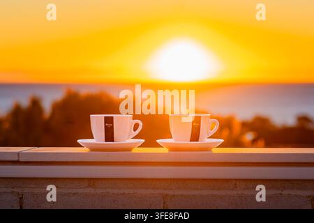 Deux tasses à espresso sur la rambarde d'un balcon face à un lever de soleil doré et vibrant sur les arbres côtiers et la mer Méditerranée. Banque D'Images