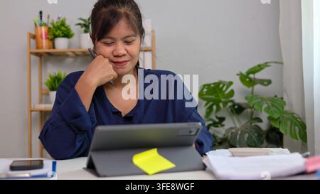 Femme d'affaires asiatique tenant un stylet et souriant tout en travaillant sur une tablette numérique au bureau à domicile. Entrepreneure asiatique regardant l'écran Banque D'Images