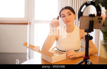 Jeune femme ouvre une boîte contenant des articles de soins de la peau tout en filmant une vidéo pour les réseaux sociaux, avec un smartphone sur un trépied et des fleurs à proximité Banque D'Images