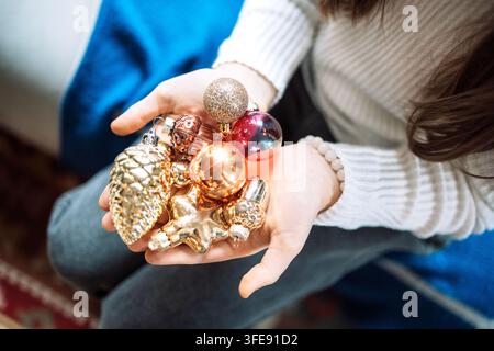 Les mains d'une femme présentent une collection d'ornements de Noël colorés, comprenant des pièces dorées et argentées brillantes, sur un fond intérieur chaleureux Banque D'Images