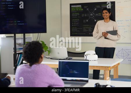 Enseignant indien debout avec une tablette dans un laboratoire tandis qu'un étudiant jeune africain code sur un ordinateur portable, espace de copie Banque D'Images