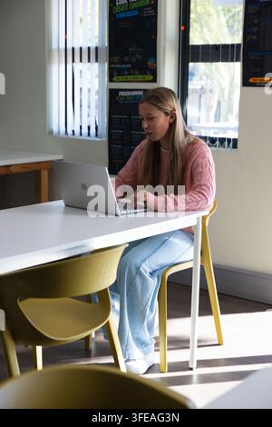 Adolescente étudiant dans une salle de classe sur une chaise jaune à une table en utilisant un ordinateur portable argenté et des affiches Banque D'Images