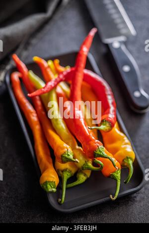 Des piments rouges, orange et jaunes vibrants disposés sur un plateau noir à côté d'un couteau de chef, mettant en valeur des produits frais, une saveur épicée et une cuisine rustique Banque D'Images