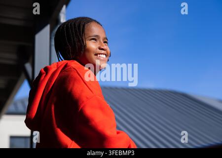 Adolescent debout sur un balcon portant un sweat à capuche rouge, regardant à droite et observant un toit en métal Banque D'Images