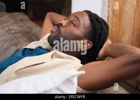 Homme africain allongé avec les mains derrière la tête sur un canapé gris à la maison, oreiller blanc, chemise beige Banque D'Images
