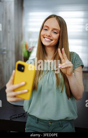 Jeune femme souriante tenant un smartphone et faisant un signe de paix pour les réseaux sociaux Banque D'Images