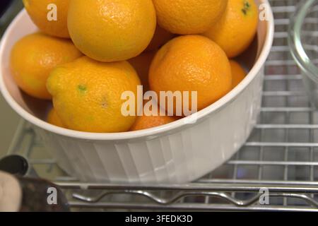 Un bol en céramique blanche rempli d'oranges fraîches et mûres repose sur une étagère en fil métallique dans une cuisine. Banque D'Images