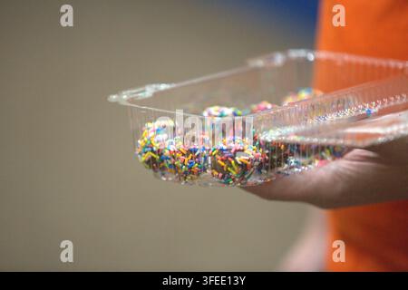 Person tenant un récipient en plastique transparent rempli de truffes au chocolat enrobées de paillettes colorées arc-en-ciel. Banque D'Images