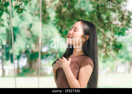 Portrait d'une jeune femme les yeux fermés posant les mains sur sa poitrine dans un parc, prenant une profonde inspiration d'air frais tout en se relaxant en plein air dans un vert paisible Banque D'Images