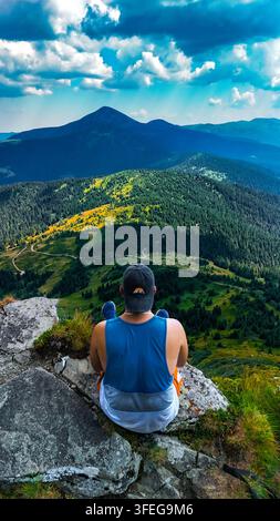 Un randonneur est assis sur une corniche rocheuse face à des crêtes montagneuses panoramiques et à un pic dominant. La scène capture une réflexion calme, l'aventure et la récompense d'un l Banque D'Images