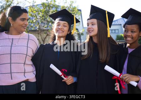 Quatre jeunes femmes souriantes debout sur la pelouse du campus, portant des robes et des toges, tenant des diplômes Banque D'Images