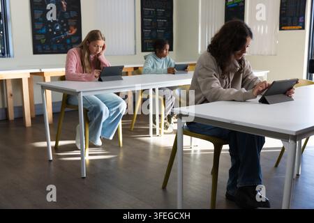 Diverses camarades de classe assis dans une salle de classe utilisant des tablettes, un jeune portant un sweat à capuche beige s'appuie sur un folio Banque D'Images