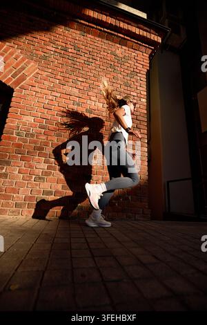 Prise de vue dynamique en contre-plongée d'une femme en forme jogging sur un trottoir urbain. Ses longs cheveux blonds volent en mouvement. Une lumière solaire forte projette une ombre dramatique sur le brique rouge Banque D'Images