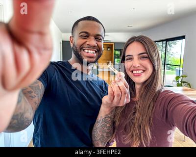 Couple divers excité célébrant la propriété d'une nouvelle maison, tenant la clé de leur propriété moderne, capturant un moment de selfie joyeux ensemble à l'intérieur de leur Banque D'Images