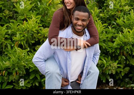 Un jeune couple partage une balade à califourchon joyeuse tout en souriant et profitant du temps à l'extérieur dans un jardin luxuriant et verdoyant, transmettant le bonheur, l'affection et la décontraction Banque D'Images