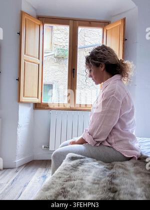 Une femme est assise sur un lit près d'une fenêtre en bois ouverte, regardant à l'extérieur dans une lumière douce du matin. Une chemise en lin décontractée, une couverture douillette et un intérieur calme transmettent une ambiance paisible Banque D'Images