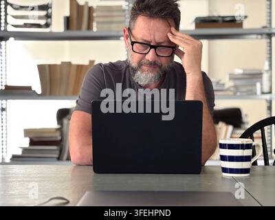 Homme d'âge moyen portant des lunettes se concentre sur un ordinateur portable dans un bureau à domicile, semblant stressé et frustré tout en tenant sa tête à côté d'un C rayé Banque D'Images