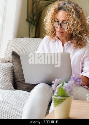 Une femme d'âge moyen aux cheveux bouclés et aux lunettes travaille sur un ordinateur portable assise sur un canapé confortable à la maison, illustrant le travail à distance, la concentration, le confort Banque D'Images