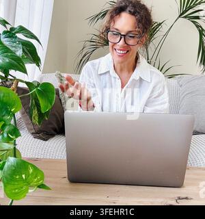 Femme portant des lunettes et une chemise blanche souriant brillamment tout en ayant un appel vidéo sur son ordinateur portable, travaillant à distance depuis son confortable bureau à domicile Banque D'Images