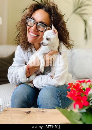 Femme souriante câlinant un petit chihuahua blanc sur un canapé, exprimant la chaleur, l'affection et le lien entre l'animal et son propriétaire dans un salon lumineux et confortable Banque D'Images