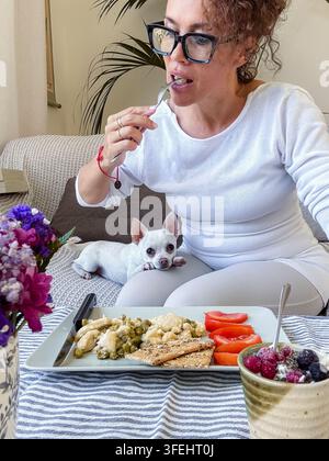 Une femme savoure un petit-déjeuner sain sur un canapé tandis qu'un petit chien blanc repose sur ses genoux, entourée de fleurs et d'une décoration d'intérieur chaleureuse, suggérant le confort et la compagnie Banque D'Images