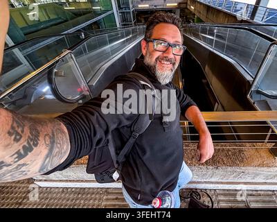 Homme souriant prenant un selfie grand angle sur une escalator urbaine dans une station de transit, portant des lunettes, un sac à dos et un bras tatoué, évoquant voyage et aventure Banque D'Images