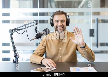 Jeune homme adulte portant des écouteurs et utilisant un microphone de studio professionnel, regardant la caméra et agitant la main tout en diffusant en direct un podcast ou une émission de radio depuis un studio de diffusion moderne Banque D'Images