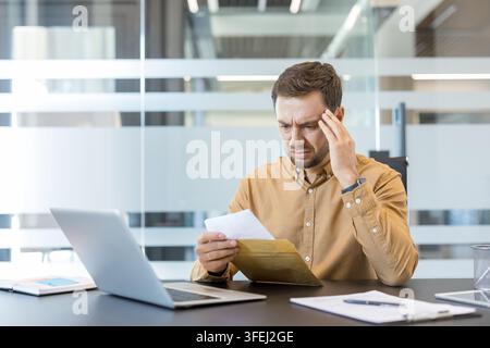 Homme d'affaires stressé au bureau tenant sa tête tout en lisant une facture inquiétante tirée d'une enveloppe, frustré et pensif face aux dettes, au risque de faillite et aux problèmes financiers Banque D'Images