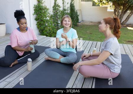 Amies féminines assises sur des tapis d'exercice noirs sur une terrasse en bois dans l'arrière-cour, tenant des bouteilles d'eau Banque D'Images