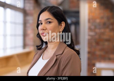Femme professionnelle posant dans un espace de travail moderne près d'une fenêtre à multiples vitres, portant un blazer marron et des créoles en or Banque D'Images