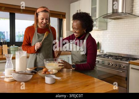 Diverses amies portant des tabliers en train de mélanger la pâte dans une cuisine domestique avec un grand bol en verre, fouet Banque D'Images